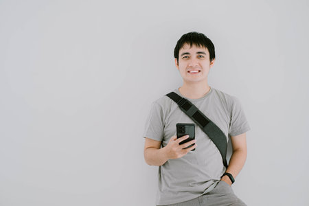 Selective focus shot of young Asian man wearing sage green casual T-Shirt, smartwatch, and sling bag is looking at camera with smile expression while holding smartphone on isolated white backgroundの写真素材
