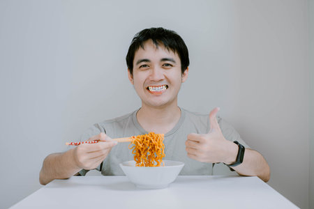 Selective focus shot of young Asian man wearing sage green casual T-Shirt and smartwatch is taking noodles from bowl using chopsticks with excited expression on isolated white backgroundの写真素材