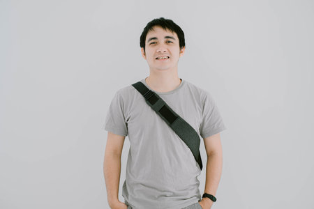 Selective focus shot of young Asian man wearing sage green T-Shirt, a smartwatch, and minimalist sling bag is looking at the camera with smile expression. Isolated white background.の写真素材