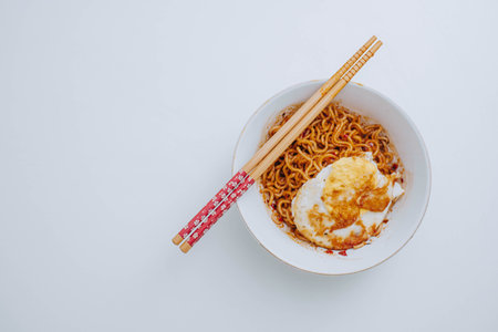 Flat lay or top view shot of a portion of stir-fried Indonesian instant noodles with egg on a white bowl with a pair of wooden chopsticks on white tableの写真素材
