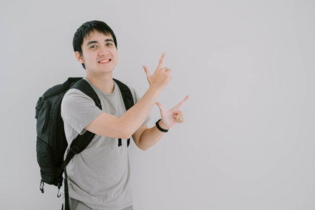 Selective focus of young Asian man wearing sage green casual T-Shirt, smartwatch and backpack with smile is pointing to the upper right side while looking at the camera on isolated white backgroundの写真素材