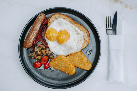 Flat lay or top view shot of a plate of hashbrown with sunny side egg, sourdough or fried potato, beef bacon, sausage, roasted mushrooms, and cherry tomatoes with knife and fork on the right sideの写真素材