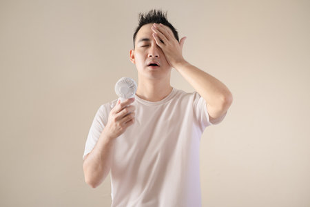 A young Asian man feel relieved when using a portable fan because of hot weatherの写真素材