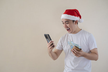 Young Asian Santa man is excited with a happy expression while looking at his smartphone and holding his wallet and money. Isolated beige background. Christmas concept.の写真素材
