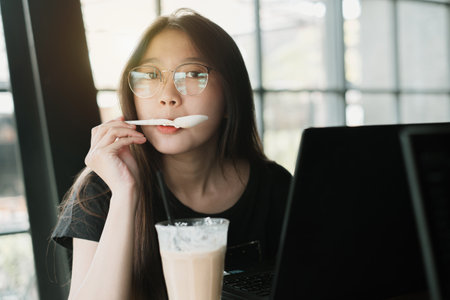 Young Asian woman is spooning toppings from a glass of coffee while looking at the camera in front...
