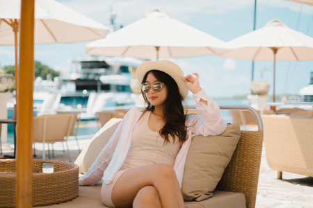Beautiful young Asian girl is sitting in the cafe near the harbor with blurred yacht and sea in the background. Wearing slim cream dress, pink and sunglasses, straw hat, necklace.の写真素材
