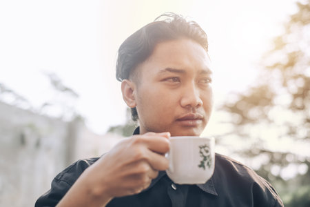 A Southeast Asian man is holding a cup of coffee during sunset in the afternoon with bokeh in the foreground and backgroundの写真素材
