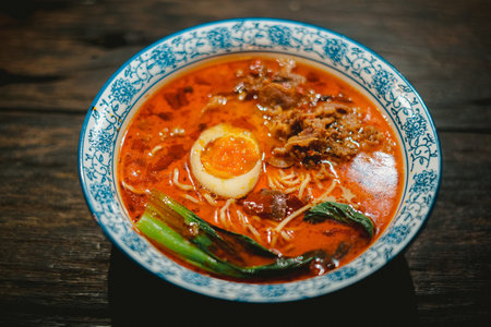 A steaming bowl of Chinese noodle soup with shredded beef meats, a hard-boiled egg, and a vegetable...