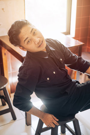 A Southeast Asian man wearing black jacket and pant is sit and pose on a wooden chair on the kopitiam indoor theme while looking at the camera with bokeh background. Portrait or vertical shot.の写真素材