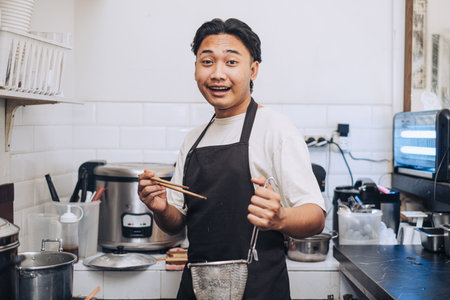 A Southeast Asian chef with an apron is drying up the noodles in the kitchen while looking at the camera with smile face expression in the afternoon with bokeh backgroundの写真素材