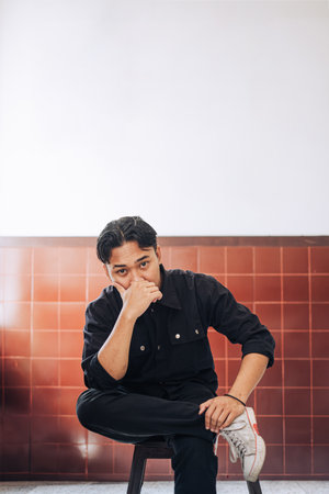 A Southeast Asian man wearing black jacket and pant is sit and pose on a wooden chair indoors while looking at the camera with bokeh background. Portrait or vertical shot.の写真素材