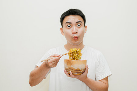 Young Asian man in white shirt picking up fried noodles on wooden bowl using chopsticks with wow or surprised face expression while looking at camera. Isolated background.の写真素材