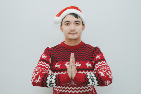 Asian man in Christmas sweater and santa hat making a prayer gesture while looking at camera on isolated white backgroundの写真素材
