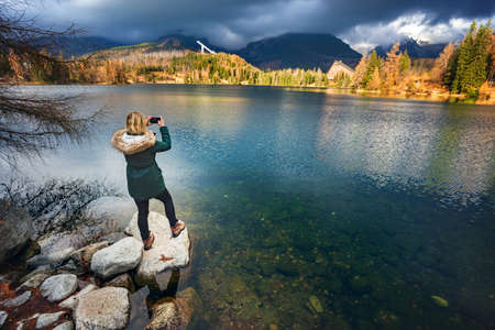 Young woman taking photo of lake in Tatra mountains in autumn. Strbske pleso in national park High Tatrasの写真素材
