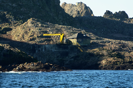 A run-down looking building with "Federal Wildlife Refuge No Trespassing No Shooting" painted on the side of it and an old crane on the Southeast Farallon Island, outside the San Francisco Bay. Sea Lions are visible on rocks near the water.のeditorial素材