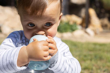 A little baby boy in a park in Spain, during a beautiful sunny day, playing outdoor. He is drinking his water bottle. He is caucasian and has blue eyes. He is wearing a sweater gray and skyblue.の写真素材