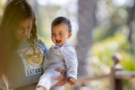 A mother having fun with her boy in a natural park in Spain during a sunny day. They are laughing and playing with each other. They are surrounded by green and big palm trees. They are having a walk.の写真素材