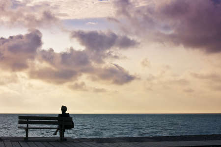 woman sitting on bench looking out to sea sunset skyの写真素材