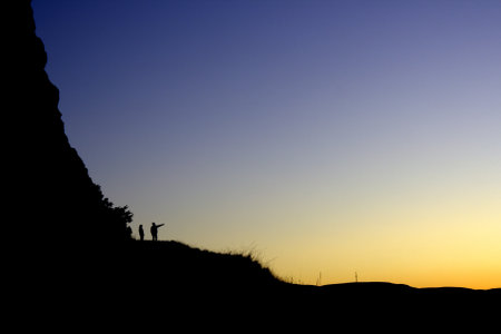 Silhouette of two people standing on top of a cliff at sunsetの写真素材