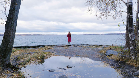 Young girl with her hair stands near the water in the cold season. Back view. Young woman standing near a lake. Calm and cozy eveningの写真素材