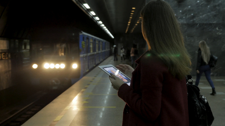Girl in red coat using smartphone or tablet at subway station and waits for the train. Woman use of cellphone and standing at city subway staton.の写真素材