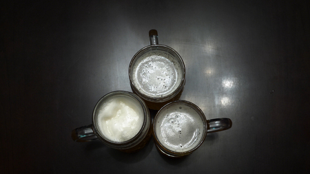 Cheers. Top view of people holding mugs with beer. Top down shot of three pint sized beer mugs on a wooden pub table. Close-up partial view of young friends clinking beer glasses. Hand holding glasses of beer clinking together at outdoor resturantの写真素材