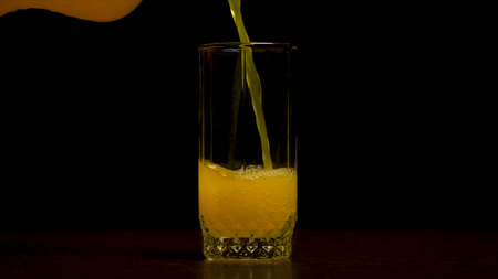 Orange soda large glass, overflowing glass of orange soda closeup with bubbles isolated on black background. Pouring Orange Drink. Slow Motion. Carbonated orange drink is poured into a glass. Pouring orange juice soda in glass in slow motion, black backgroundの写真素材