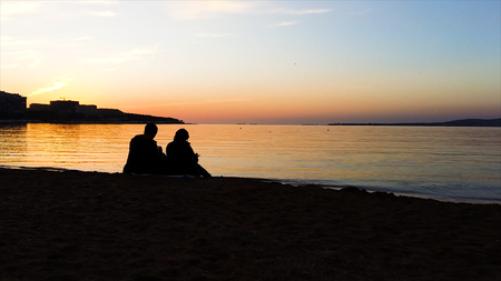 Silhouette of man and woman watching the sunset. Silhouette of the couple enjoying the sunset on the beach. Young couple sitting on a bench at sunset. There is a flying bird, silhouetteの写真素材