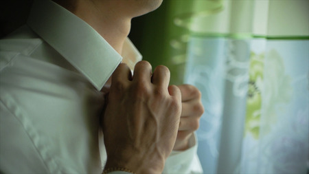 Man buttons up his white shirt standing in the front of a bright window. Clip. People, business, fashion and clothing concept - close up of man dressing up and fastening buttons on shirt at home.の写真素材