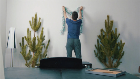 Man decorates the wall with Christmas decorations, decorate the tree. Young man hanging Christmas...