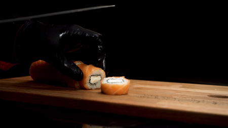 Hands in black gloves cut Philadelphia roll with sharp knife on wooden board, isolated on black background. Salmon and cream cheese roll cut into small pieces by sushi chef, foodporn concept.の写真素材