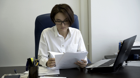 Young and beautiful businesswoman with short, dark hair in white shirt is tired from work in the office. Young office lady looks concentrated and tired looking through documents, typing on computer.の写真素材
