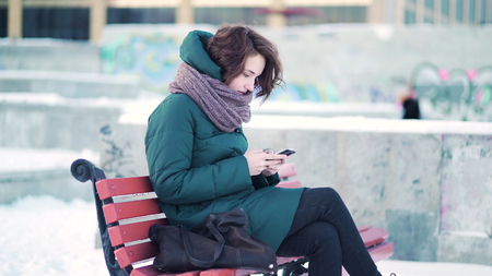 Happy woman texting on a smart phone while sitting on a bench in winter city against stone blocks with graffiti background. Lovely brunette browsing on her mobile phone while resting on a bench.の写真素材
