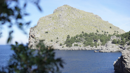 Mountain landscapes covered with green vegetation against blue sky. Art. Rocky cliff covered with trees and shrubs on shore with blue water at foot on clear summer dayの写真素材