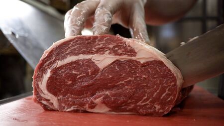 Butcher in gloves cuts carcass of red meat. Action. Close-up of professional knife cutting huge piece of meat in butcher shop.の写真素材