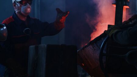 Workers poured molten metal at plant. Stock footage. Workers in form and helmets control process of pouring molten metal from boiler at metallurgical plantの写真素材
