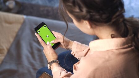 Pretty woman with brown hair sitting at home in pink shirt and sliding on her smartphone at the chroma key green screen. Stock footage. Smartphone new technology conceptの写真素材