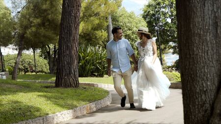 Newlyweds run in park in summer. Action. Beautiful loving couple romantically run away holding hands in park. Wife in white dress holding her husbands hand illuminated by sunlightの写真素材