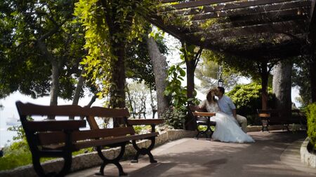 Couple sitting on bench in beautiful gazebo. Action. Beautiful couple sitting in gazebo in honeymoon. Husband kisses his precious wife on cheek. Sweet and romantic coupleの写真素材