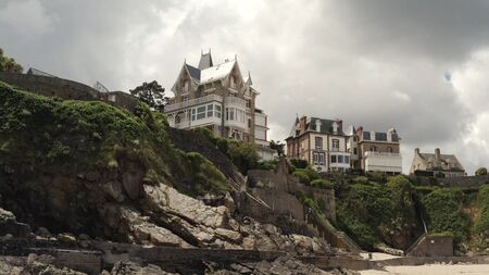 View from the riverside on the awesome fashionable cottages standing on the top of slope near the river in summer against grey cloudy sky. Action. Beautiful European architectureの写真素材