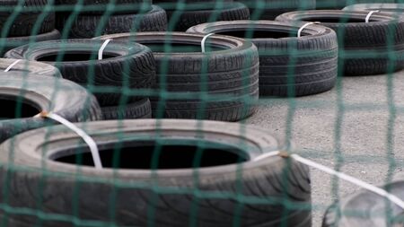 Safety barrier made of old black tires lying on a concrete pavement behind the green mesh fence. Media. Many fastened car tires used for making a track barriers.の写真素材