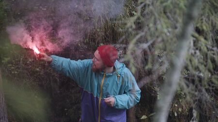 Young man holds up red smoke flare to signal for help in the middle of green wild nature. Hipster...