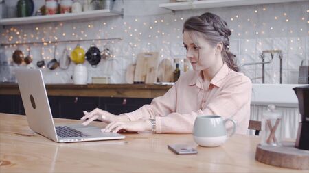 Young attractive woman working on laptop at home in kitchen. Stock footage. Beautiful woman carefully working on laptop in freelance mode and background of comfortable kitchenの写真素材
