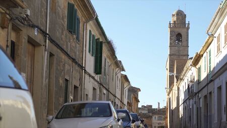 Narrow calm city street with parked cars along the old houses on blue cloudy sky background. Someone...