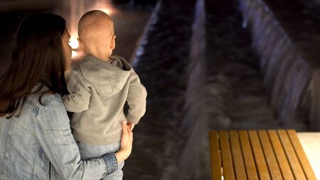 Rear view of young mother with her little son standing near the fountain in the late evening. Stock footage. Mom and her boy looking at the fountain.の写真素材