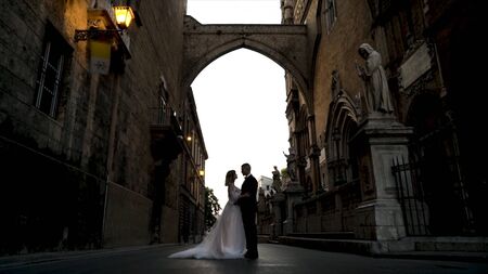 Beautiful couple of newlyweds are standing on old street. Action. Newlyweds in wedding dresses stand on background of dark old streetの写真素材