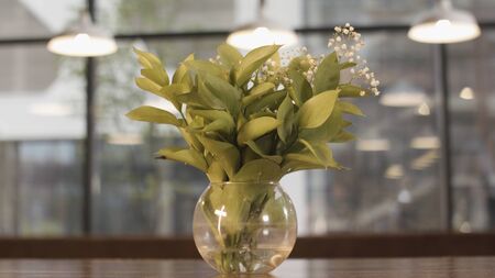 Gentle lilly of valley flowers in glass vase standing on wooden table on blurred background with windows and lamps. Spring may lily bouquet, decoration of room.の写真素材