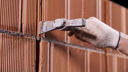 Close up of a worker holding metal hammer and making a hole inside the red ceramic block wall. Stage...