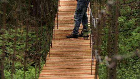 Side view of a man standing on the wooden hanging bridge and smoking a pipe while watching in the distance. Hiker having a rest and enjoying summer forest.の写真素材