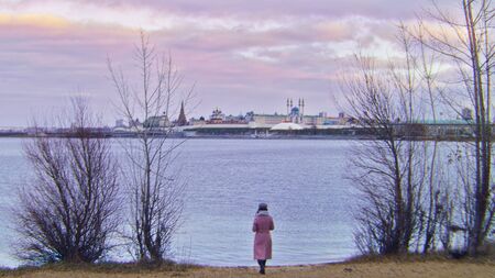 Woman walking on beach on background of old city in autumn. Stock footage. Back view of attractive young woman walking on autumn beach on background of water and city with cloudy skyの写真素材